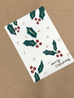 a christmas card with holly and berries on white paper, sitting on a brown surface