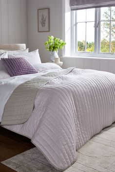 a bed with white and purple comforters in a bedroom next to a window,