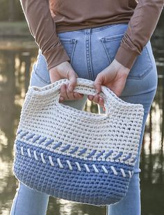 a woman is holding a crocheted bag in her hands while standing next to the water