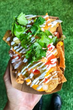 a hand holding a paper container filled with nachos covered in cheese and toppings