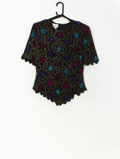 Vintage sequin top in black with floral pattern by Laurence Kazar. This beautiful floral sequin and beaded top features bright blue, pink and purple flowers, short sleeves, shoulder pads, a quarter zip and hook and eye closure on the back. Made from a silk and a polyester lining, this top is the perfect party piece! Our recommended size: Medium Label says: Large Condition: Very good* Material: 100% silk Lining material: 100% polyester  Measurements in inches: Pit to pit: 19 Shoulders: 16 Sleeve: Fitted Sequin T-shirt For Summer, Multicolor Sequined Tops For Party Season, Glamorous Multicolor Tops For Party Season, Spring Party Multicolor T-shirt, Glamorous Multicolor Top For Party Season, Purple Short Sleeve Top For Party, Glamorous Multicolor Embellished Tops, Fitted Purple Sequin Top, Fitted Sequined T-shirt For Party Season