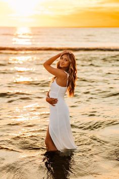 a woman standing in the water at sunset wearing a white dress and holding her hair back