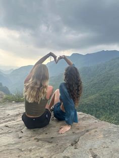 two young women sitting on top of a mountain making a heart shape with their hands