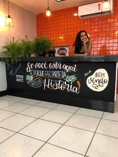 a woman sitting behind a counter with plants in the corner and writing on the wall