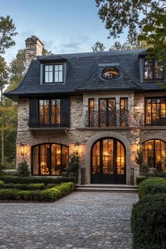 a large stone house with lots of windows and lights on the front door, surrounded by hedges