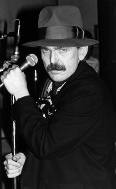 a man wearing a hat and holding a microphone in his right hand while looking at the camera