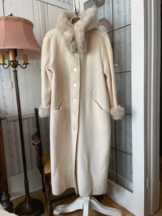 This is an iconic Canadian coat: Linda Lundstrom's La Parka! This one is made from 100 per cent virgin wool in ivory/off white, beautifully trimmed with faux fur in matching light soft beige. The faux fur trim around the collar can be removed and reattached using the zipper, and the trim around the cuffs can be attached and reattached with Velcro. The coat has the original decorative mother of pearl buttons, decorative stitching around seams and edges, two front pockets and a large hood. Marked size small. The measurements, taken with the coat lying flat, are: shoulder to shoulder, 17 inches; armpit to armpit, 22 inches; sleeves, 24 inches; length, 53 inches; bottom edge, 30 inches. In very good condition. Cream Fitted Long Wool Coat, Fitted Long Cream Wool Coat, Elegant Cream Fur Coat With Long Sleeves, Elegant Long Sleeve Cream Fur Coat, Classic Cream Wool Sweater Coat, Classic Fitted Cream Wool Coat, Classic Cream Sweater Coat For Winter, Classic Beige Fur Coat With Faux Fur Trim, Elegant Cream Fur Coat With Faux Fur Trim