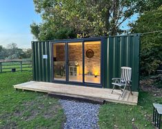 a small green building with sliding glass doors and chairs on the outside, surrounded by grass