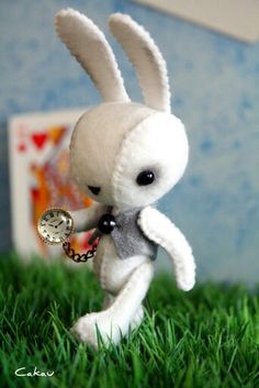 a white stuffed rabbit holding a clock in its hand on some grass with a blue wall behind it