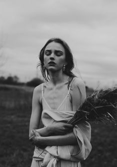 a woman standing in a field with her arms around her body and looking at the camera