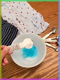 a person holding a spoon over a bowl with blue liquid in it and clothes on the table