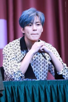 a young man with blue hair sitting at a table in front of a red curtain