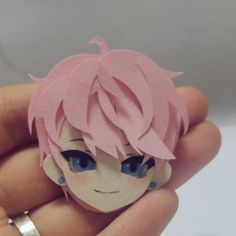 a hand holding a small paper doll with pink hair and blue eyes on it's face