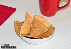 a white bowl filled with tortilla chips next to a red mug on a table