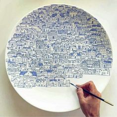 a person is drawing on a white plate with blue buildings in the background and one hand holding a pencil