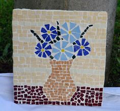 a mosaic vase with blue flowers in it sitting on a white table cloth next to a tree