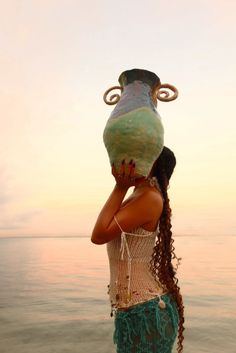 a woman standing in front of the ocean with a vase on her head and long hair