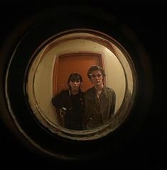 two young men standing in front of a door looking at the camera through a round window