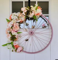 a bicycle wheel with flowers on it hanging from the side of a door, in front of a window