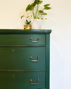 a green dresser with gold handles and plants on top, against a white wall behind it