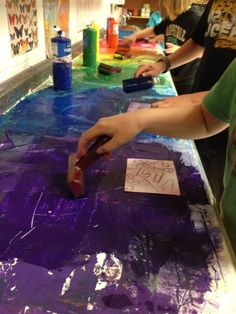 two people are painting on a table with purple paint and other items in the background