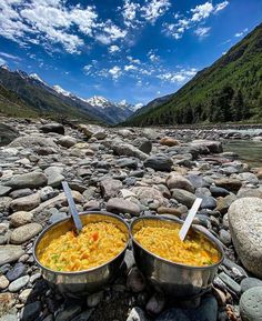 two metal bowls filled with food sitting on top of a rocky river bank next to mountains