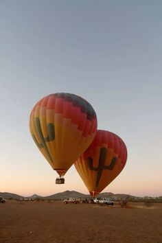 two hot air balloons flying in the sky over a desert area with vehicles parked nearby