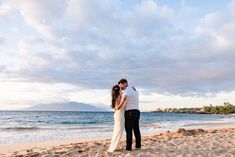 a man and woman standing on top of a sandy beach next to the ocean under a cloudy sky