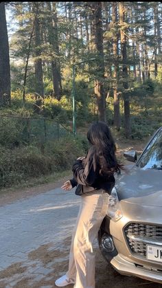 a woman standing next to a silver car on a dirt road in front of trees