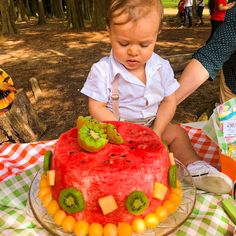 a young child sitting in front of a cake with fruit on it and people standing around