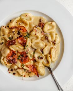 a white plate topped with pasta covered in sauce and bacon next to a fork on top of a table