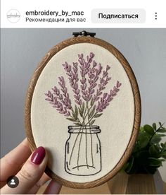 a person holding up a small embroidery project with purple flowers in a mason jar on it