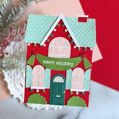 a red house shaped christmas ornament hanging from a silver tree with a pink and green background