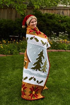 a woman is standing in the grass with a christmas tree blanket on her shoulders and wearing a knitted hat