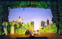 children are sitting on the ground in front of a stage with green plants and rocks