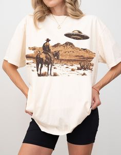 Hey y'all! If you're lookin' to add a little Southern charm and a touch of the unexpected to your wardrobe, this Vintage Style Space Cowboy T-Shirt is just the thing! In the perfect shade of sand, this Comfort Colors C717 shirt features a unique design of a cowboy on his horse, gazing up at a UFO in the sky. You're gonna love the vintage space cowboy design, full of character and whimsy. The sand color goes with everything in your closet, and the Comfort Colors fabric is super soft and durable. Pop Culture Screen Print Tops For Fall, Unisex Graphic Tee For Fall, Pop Culture Graphic Print Tops For Fall, Pop Culture Screen Print Tops With Relaxed Fit, Pop Culture Graphic Print Tops In Relaxed Fit, Vintage Tops With Character Print In Relaxed Fit, Relaxed Fit Graphic Print Tops For Pop Culture, Vintage Tops With Character Print And Relaxed Fit, Unisex Graphic Print Top For Halloween