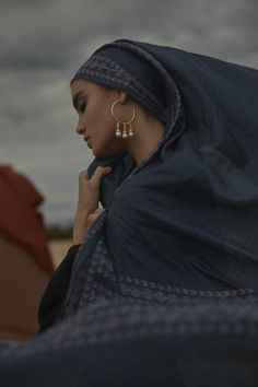 a woman wearing a blue shawl and gold hoop earrings standing in front of a tent