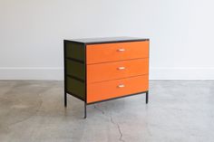 an orange and black chest of drawers in a white walled room with concrete flooring