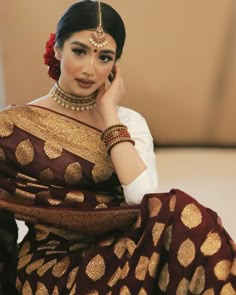 a woman in a maroon and gold sari with jewelry on her head sitting down