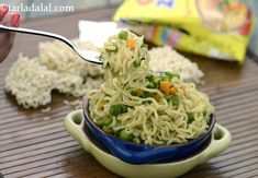 someone is holding a spoon full of noodles and vegetables in a blue bowl on a wooden table