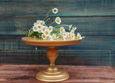 daisies in a gold pedestal on a wooden table with wood planks behind it