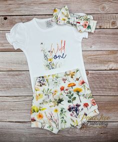 a white shirt and floral shorts with the words wild one printed on it, sitting next to a wooden background