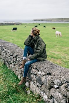 a woman sitting on a stone wall with cows grazing in the field behind her and looking off into the distance