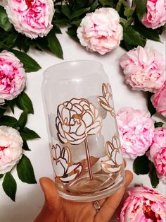 a person holding up a glass with flowers on it in front of pink peonies