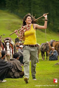 a woman holding a stick in her right hand while walking through a field with horses