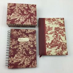 three notebooks are sitting next to each other on a white surface, one is red and the other is beige