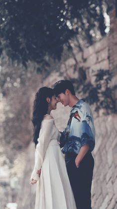 a man and woman standing next to each other in front of a stone wall with trees