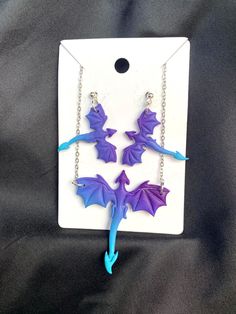 three purple and blue dragon dangles hang from chain earrings on a black cloth background