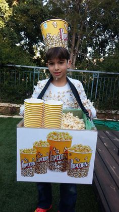 a young boy wearing a popcorn costume and holding a cardboard box with cups on it