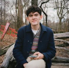 a young man sitting on a wooden bench in the woods holding a cell phone and looking at the camera